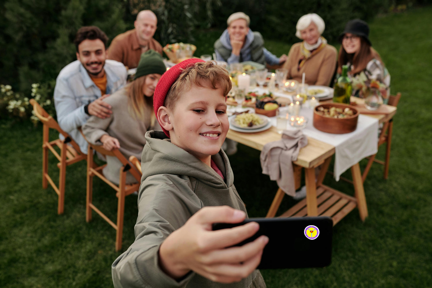 Child taking a selfie at a family gathering outdoors with a table full of food.